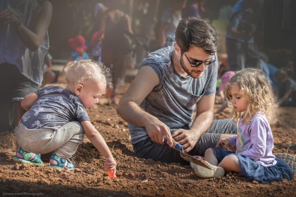 אב בין שני ילדיו יושבים על האדמה ושותלים צמחים