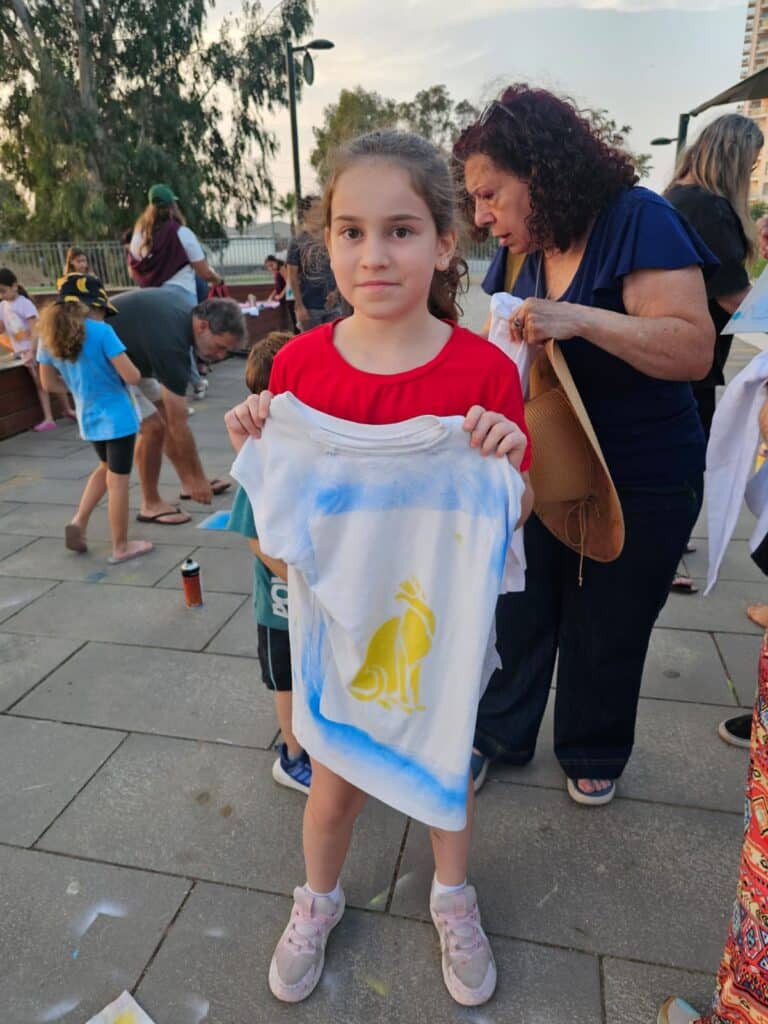 ילדה מחזיקה בחולצה לבנה עליה ציירה בספריי צבעוני עם סטנסיל