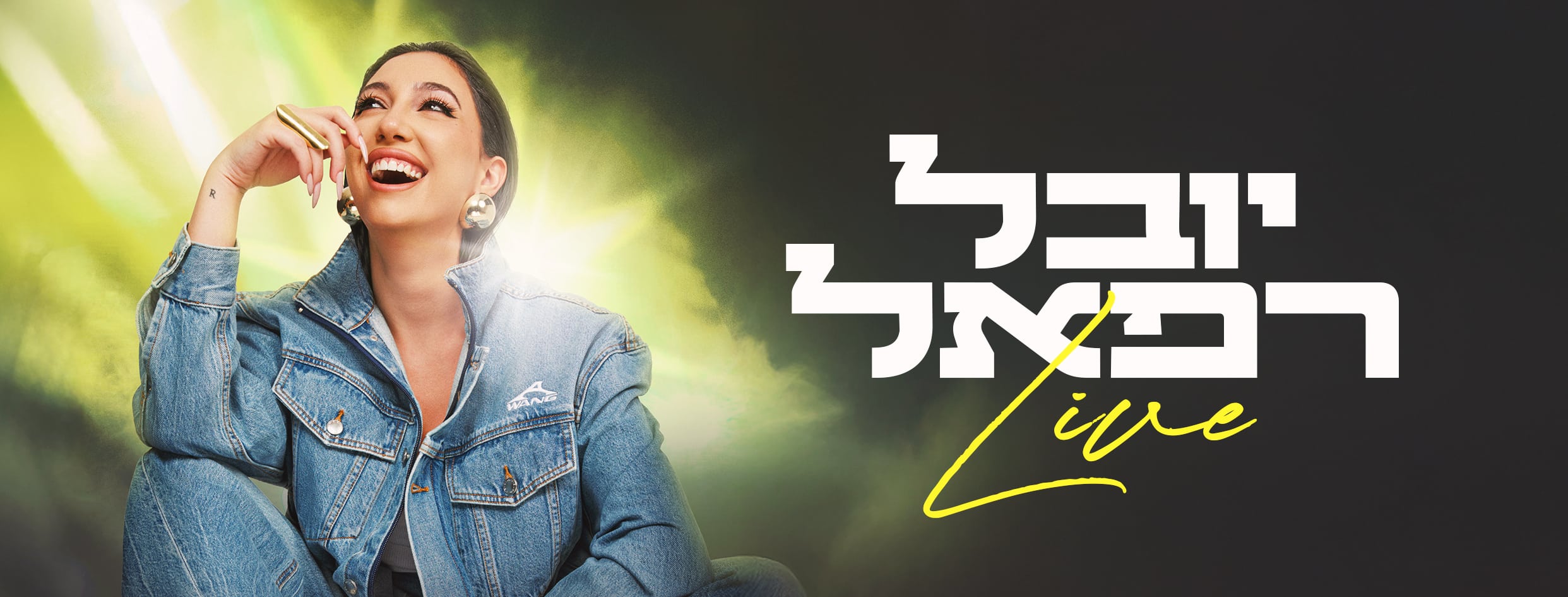 יובל רפאל בחליפת ג'ינס יושבת בישיבה מזרחית ומאחוריה אור ירוק