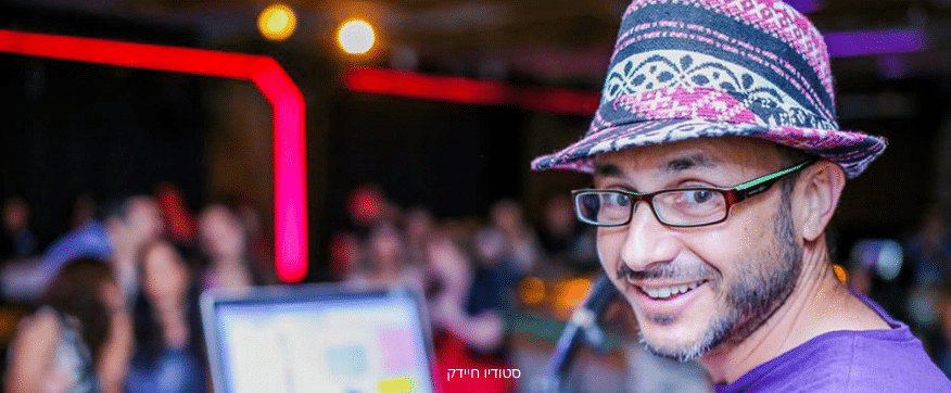 נגן בכובע ומשקפיים מחייך למצלמה על רקע קהל