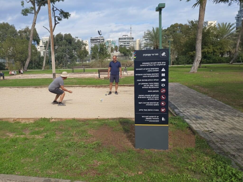 שלט ניצב בכניסה למגרשי פטאנק ושני שחקנים משחקים במגרש